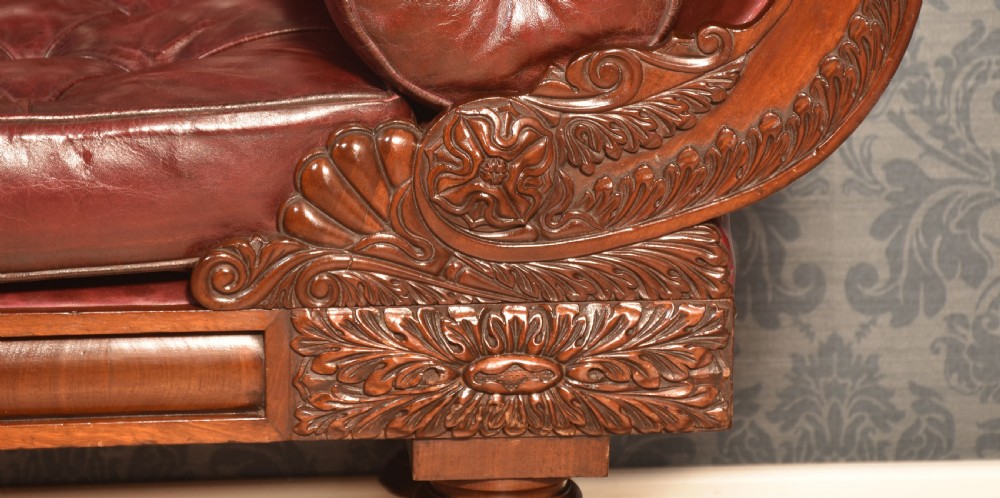 An early 19th-century deep-buttoned burgundy leather scroll-end settee with a carved mahogany frame, bolster cushions, and turned legs on brass castors. View VIII