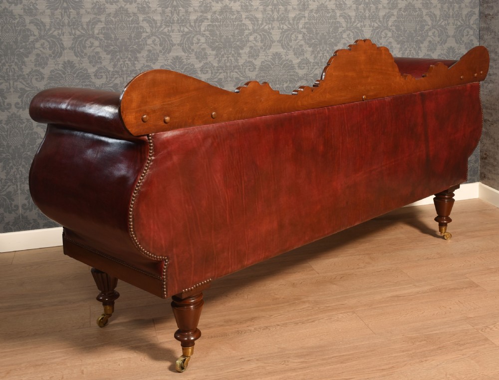 An early 19th-century deep-buttoned burgundy leather scroll-end settee with a carved mahogany frame, bolster cushions, and turned legs on brass castors. View IX