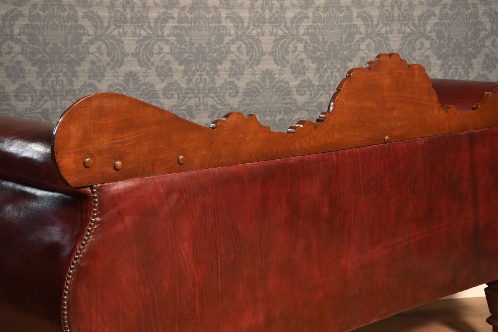 An early 19th-century deep-buttoned burgundy leather scroll-end settee with a carved mahogany frame, bolster cushions, and turned legs on brass castors. View X