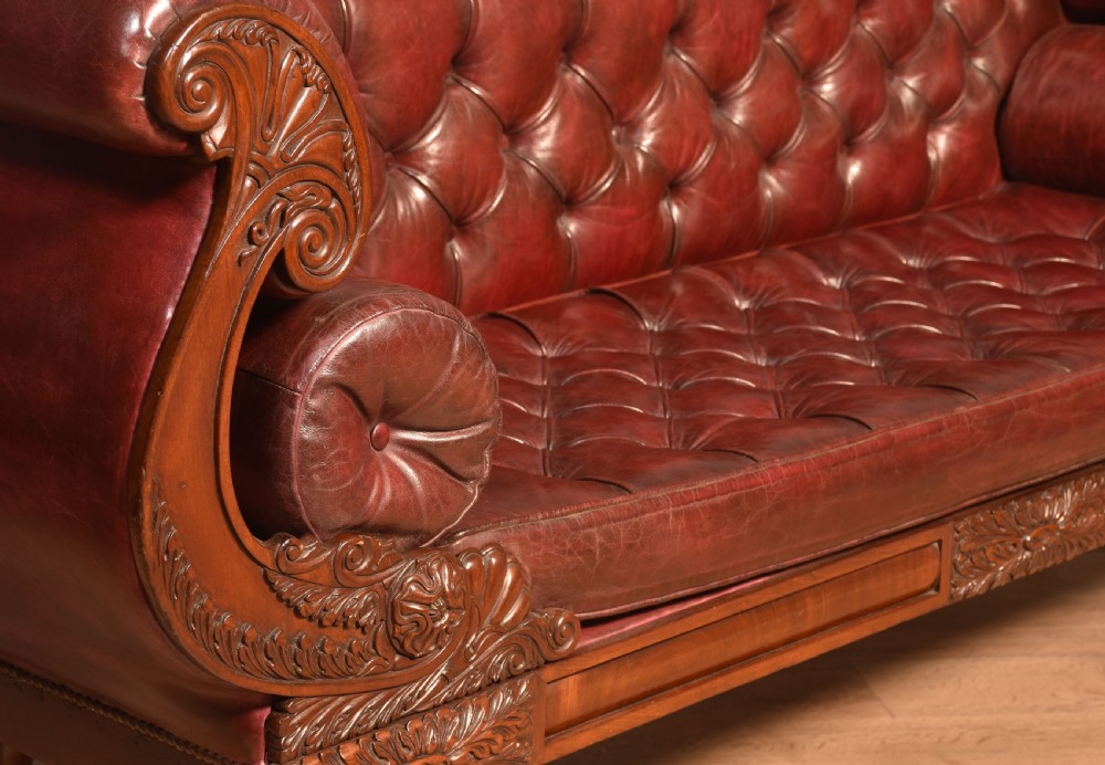 An early 19th-century deep-buttoned burgundy leather scroll-end settee with a carved mahogany frame, bolster cushions, and turned legs on brass castors. View XIII