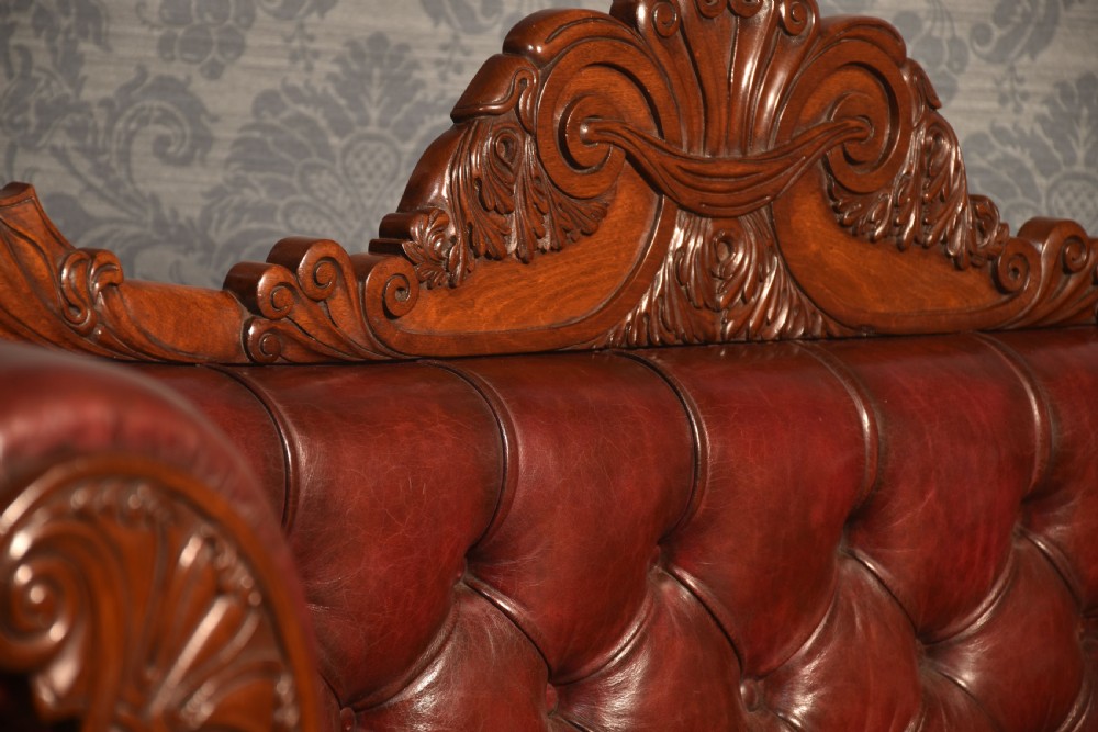 An early 19th-century deep-buttoned burgundy leather scroll-end settee with a carved mahogany frame, bolster cushions, and turned legs on brass castors. View XIV