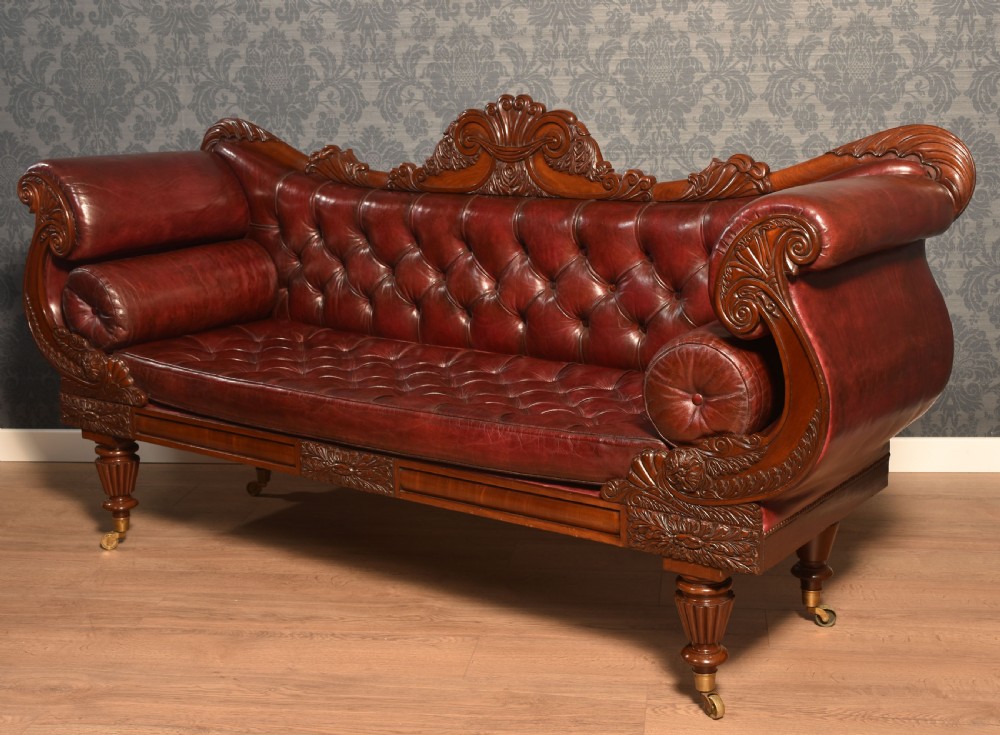 An early 19th-century deep-buttoned burgundy leather scroll-end settee with a carved mahogany frame, bolster cushions, and turned legs on brass castors. View XV