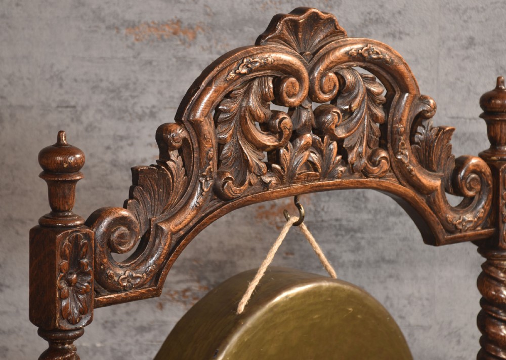 A free-standing carved dark oak dinner gong featuring barley twist columns, intricate scroll cresting, a suspended circular brass disc, and a leather striker. View VI