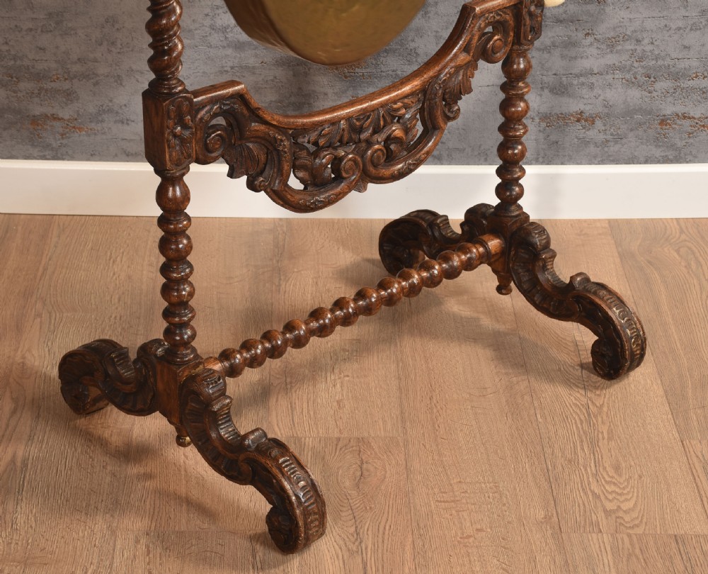 A free-standing carved dark oak dinner gong featuring barley twist columns, intricate scroll cresting, a suspended circular brass disc, and a leather striker. View VII