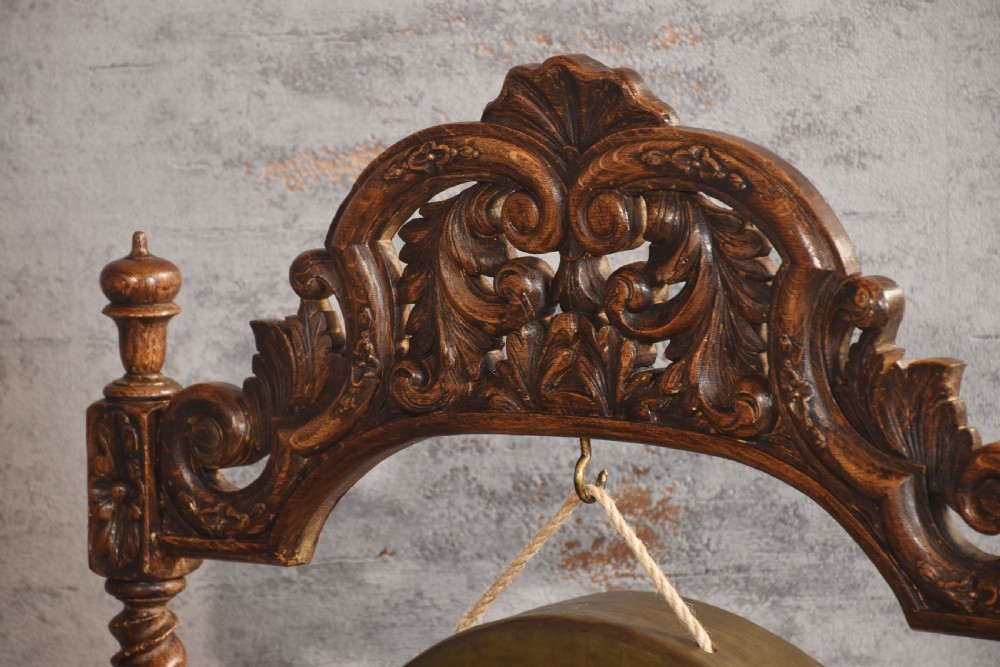 A free-standing carved dark oak dinner gong featuring barley twist columns, intricate scroll cresting, a suspended circular brass disc, and a leather striker. View X