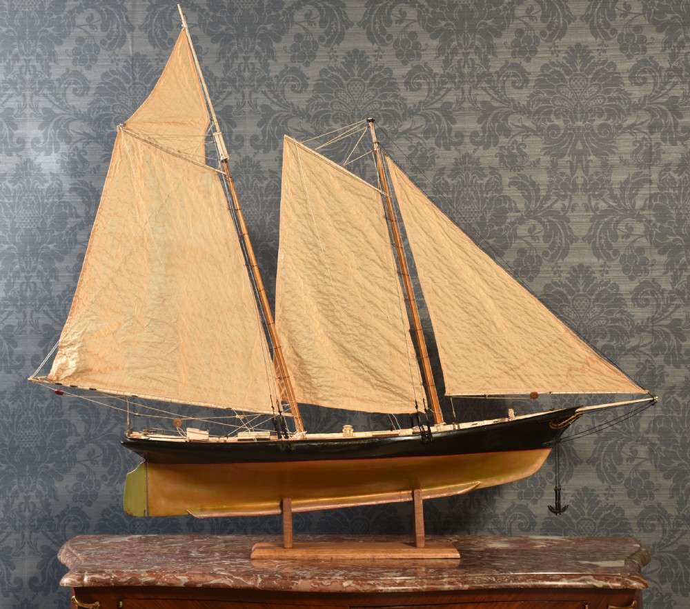 A detailed wooden model of a twin-masted racing yacht with cream canvas sails, a painted black upper hull, and a natural wood display stand. View III