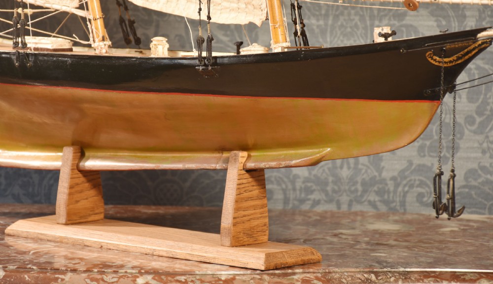 A detailed wooden model of a twin-masted racing yacht with cream canvas sails, a painted black upper hull, and a natural wood display stand. View IV