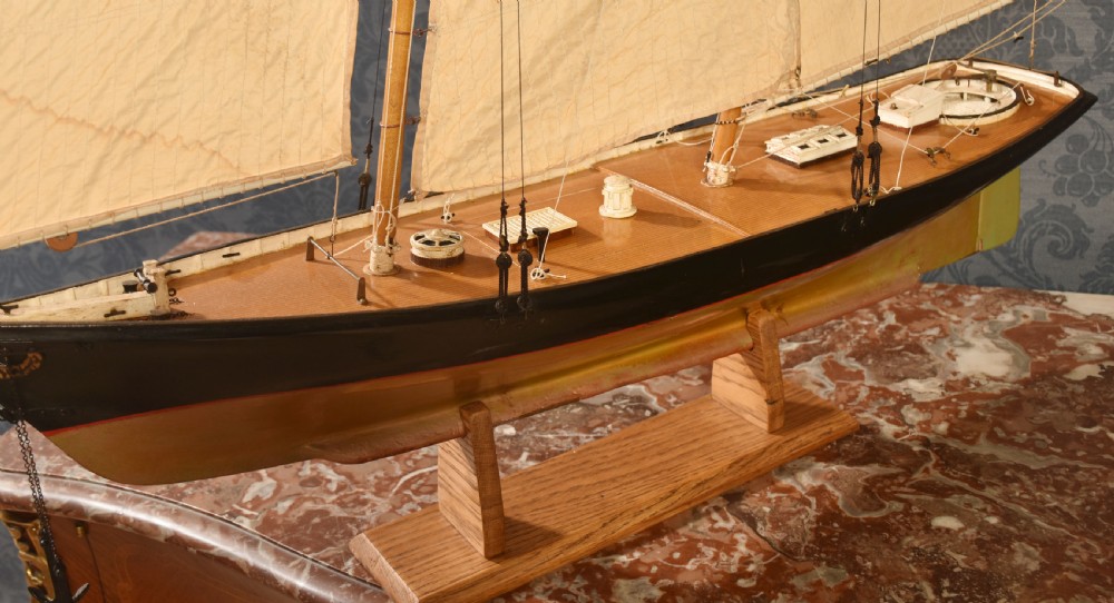 A detailed wooden model of a twin-masted racing yacht with cream canvas sails, a painted black upper hull, and a natural wood display stand. View V