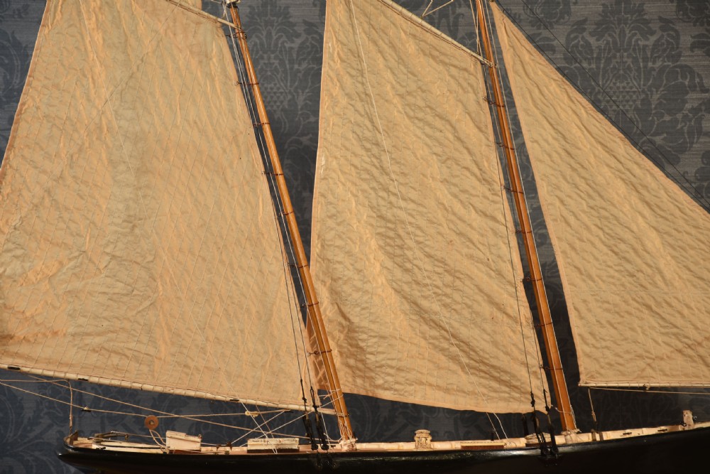 A detailed wooden model of a twin-masted racing yacht with cream canvas sails, a painted black upper hull, and a natural wood display stand. View VII