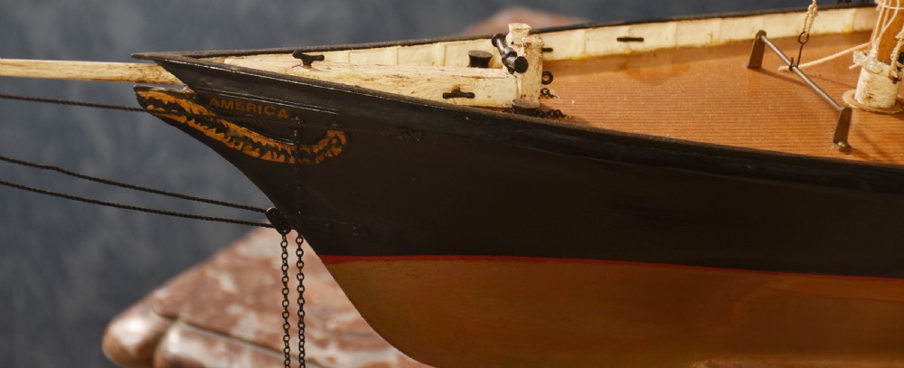 A detailed wooden model of a twin-masted racing yacht with cream canvas sails, a painted black upper hull, and a natural wood display stand. View VIII