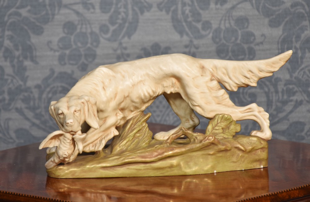 An antique Royal Dux matt porcelain figurine depicting a setter dog holding a game bird on a foliage base.