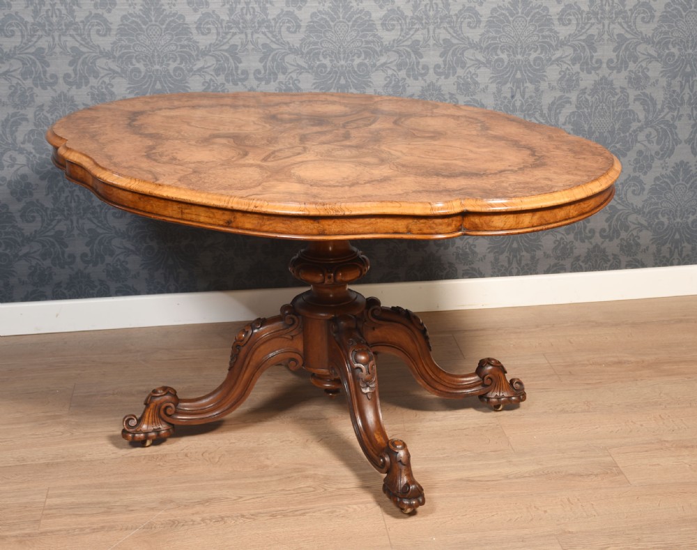 A Victorian oval burr walnut centre table with a shaped edge, turned central pedestal, and four ornate scroll-carved legs on brass castors. View III