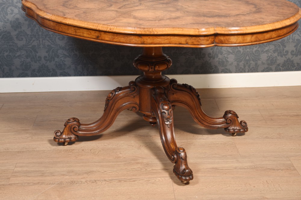 A Victorian oval burr walnut centre table with a shaped edge, turned central pedestal, and four ornate scroll-carved legs on brass castors. View V