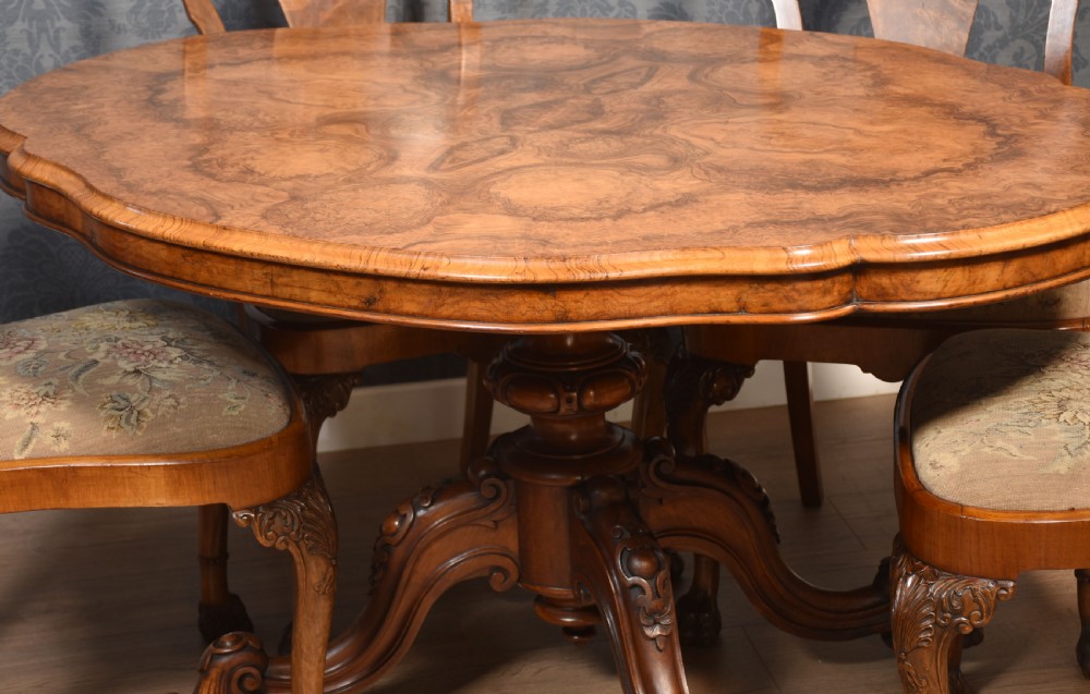 A Victorian oval burr walnut centre table with a shaped edge, turned central pedestal, and four ornate scroll-carved legs on brass castors. View VI