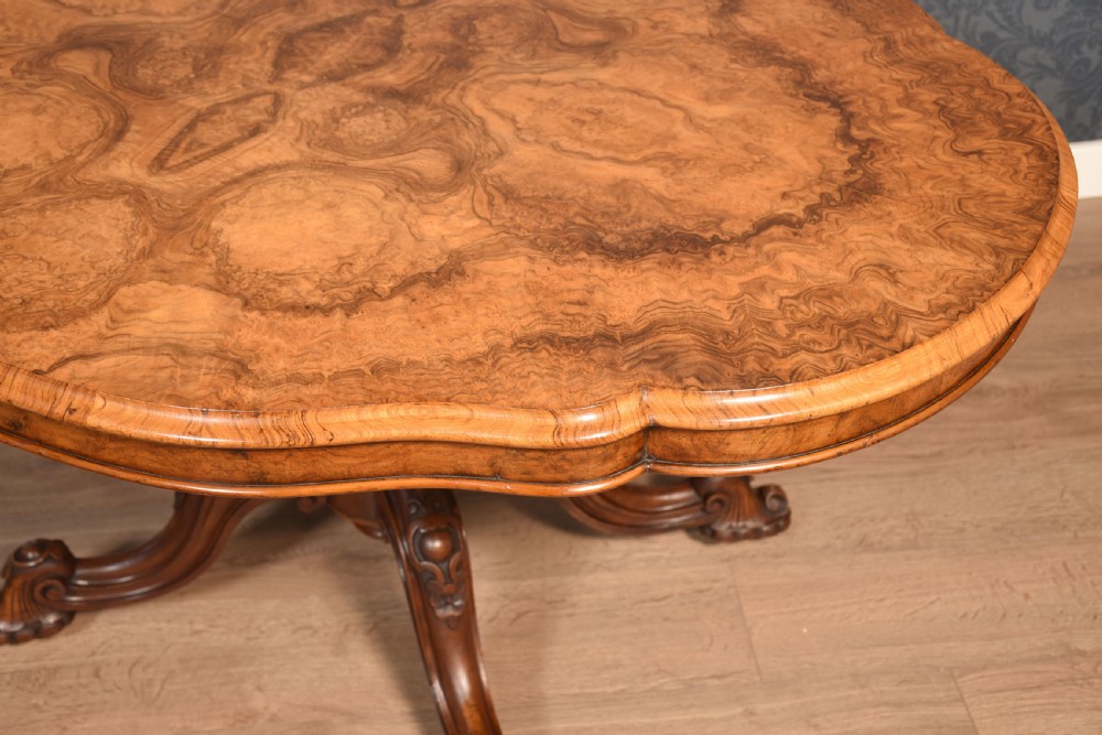 A Victorian oval burr walnut centre table with a shaped edge, turned central pedestal, and four ornate scroll-carved legs on brass castors. View VIII