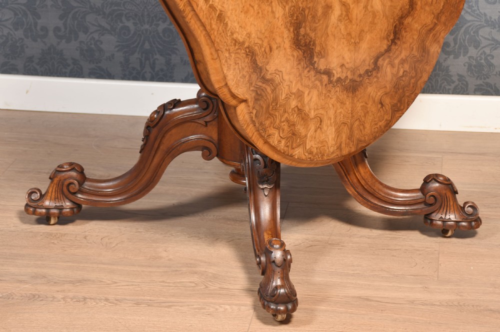 A Victorian oval burr walnut centre table with a shaped edge, turned central pedestal, and four ornate scroll-carved legs on brass castors. View XI