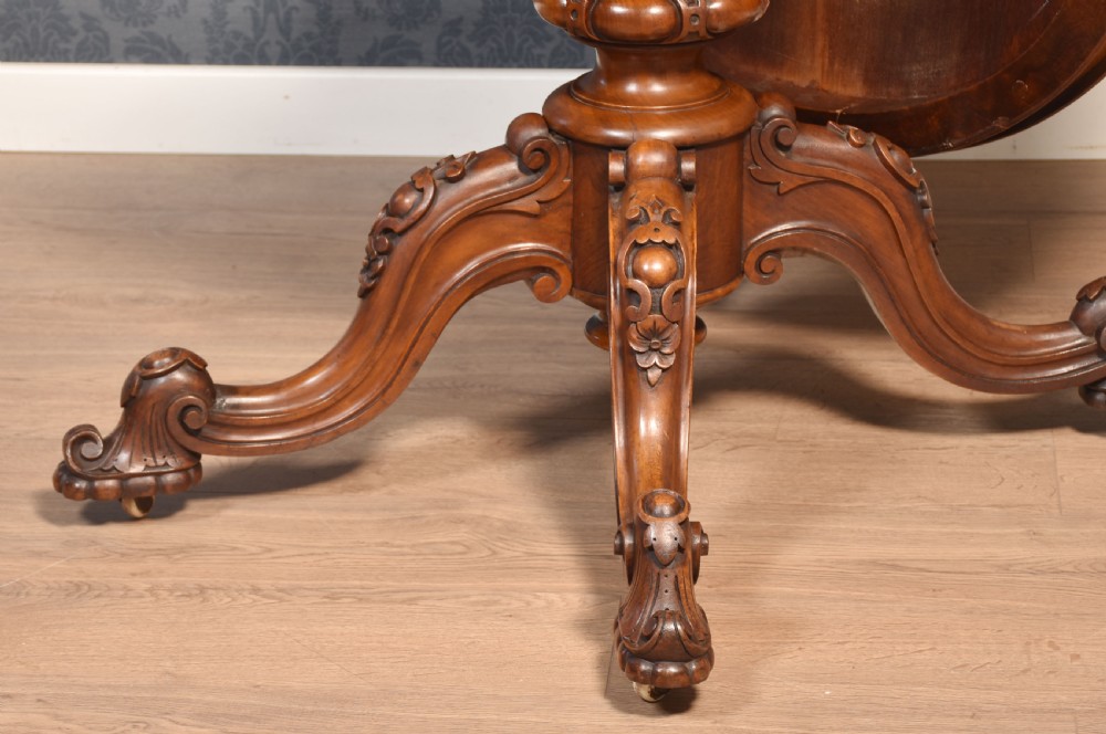 A Victorian oval burr walnut centre table with a shaped edge, turned central pedestal, and four ornate scroll-carved legs on brass castors. View XIII