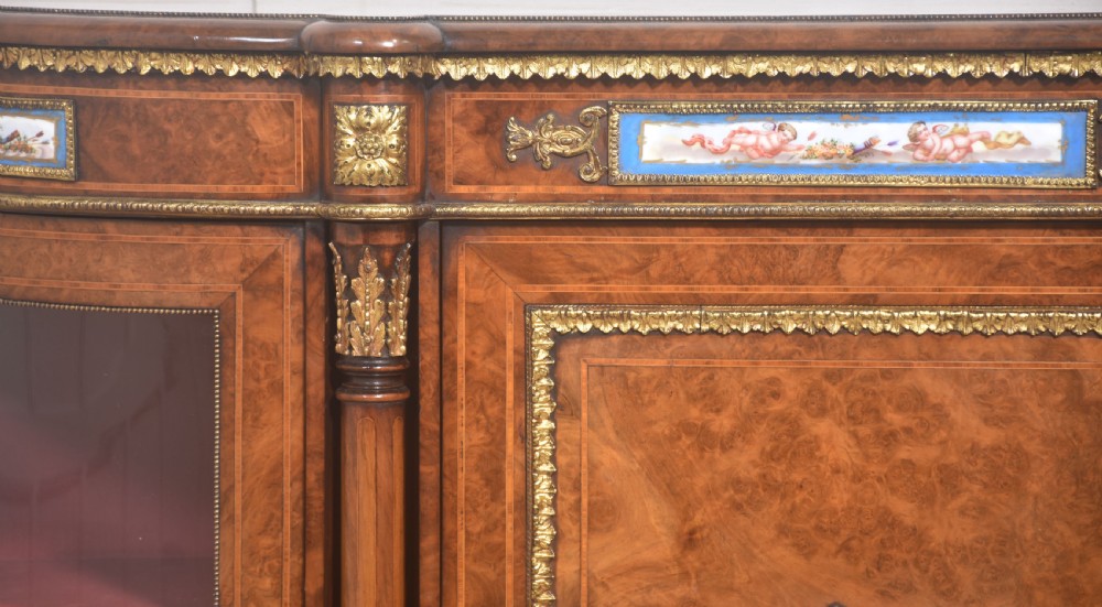 A Victorian burr walnut credenza featuring a central solid cupboard door with a porcelain plaque flanked by two bowed glazed side doors and gilt metal mounts. View VII