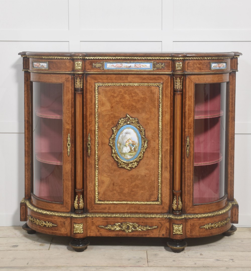 A Victorian burr walnut credenza featuring a central solid cupboard door with a porcelain plaque flanked by two bowed glazed side doors and gilt metal mounts. View IX