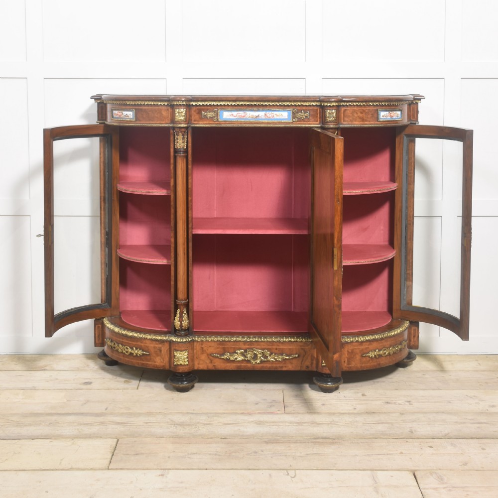 A Victorian burr walnut credenza featuring a central solid cupboard door with a porcelain plaque flanked by two bowed glazed side doors and gilt metal mounts. View XI