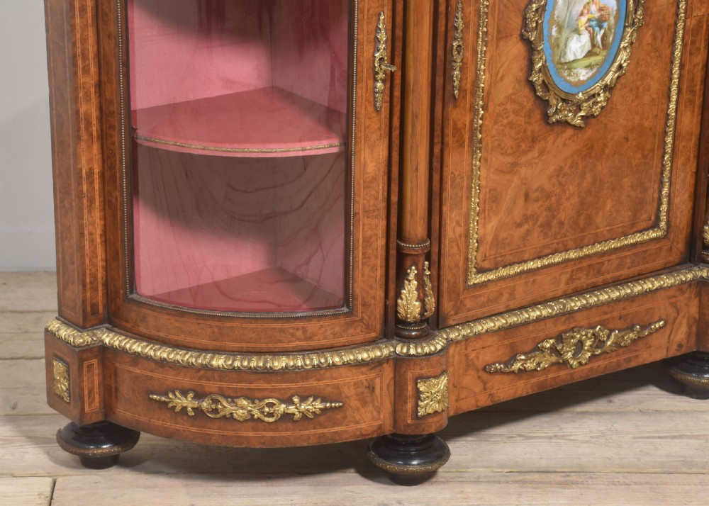 A Victorian burr walnut credenza featuring a central solid cupboard door with a porcelain plaque flanked by two bowed glazed side doors and gilt metal mounts. View XV