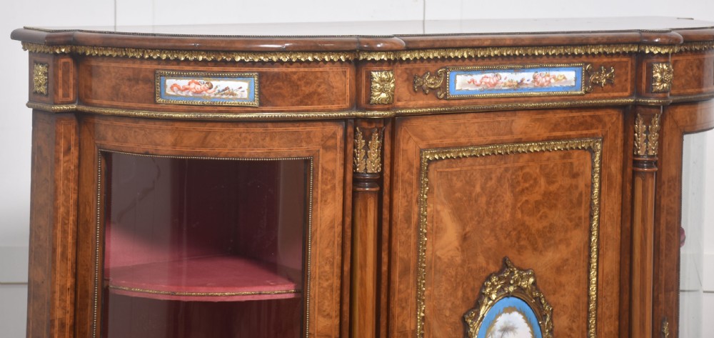 A Victorian burr walnut credenza featuring a central solid cupboard door with a porcelain plaque flanked by two bowed glazed side doors and gilt metal mounts. View XVI