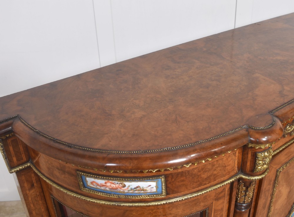A Victorian burr walnut credenza featuring a central solid cupboard door with a porcelain plaque flanked by two bowed glazed side doors and gilt metal mounts. View XVIII
