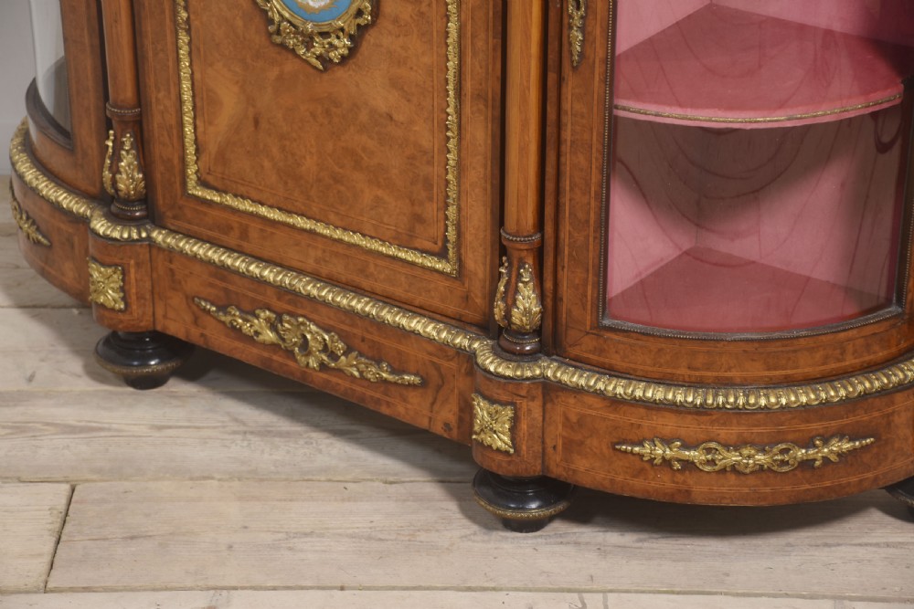 A Victorian burr walnut credenza featuring a central solid cupboard door with a porcelain plaque flanked by two bowed glazed side doors and gilt metal mounts. View XX