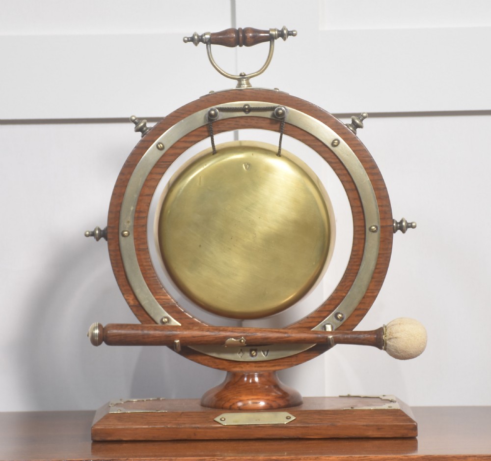 An antique oak ship wheel frame holding a central suspended brass dinner gong with an accompanying wooden striker resting on brass brackets on a rectangular base.