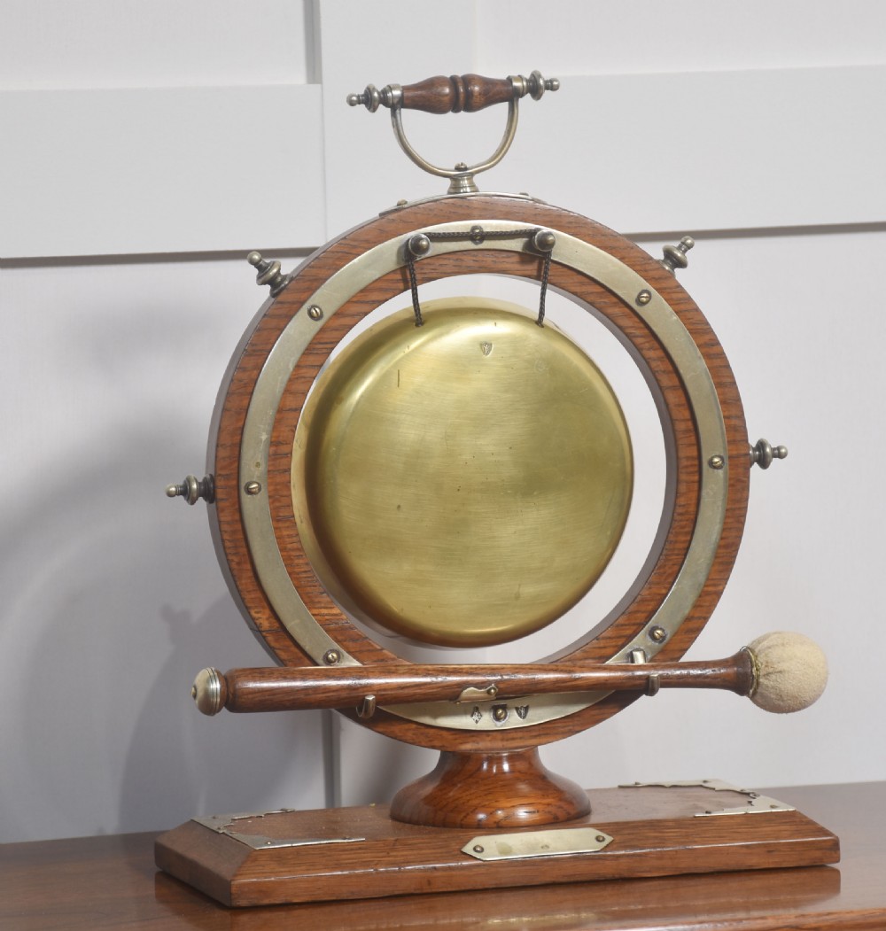 An antique oak ship wheel frame holding a central suspended brass dinner gong with an accompanying wooden striker resting on brass brackets on a rectangular base. View III
