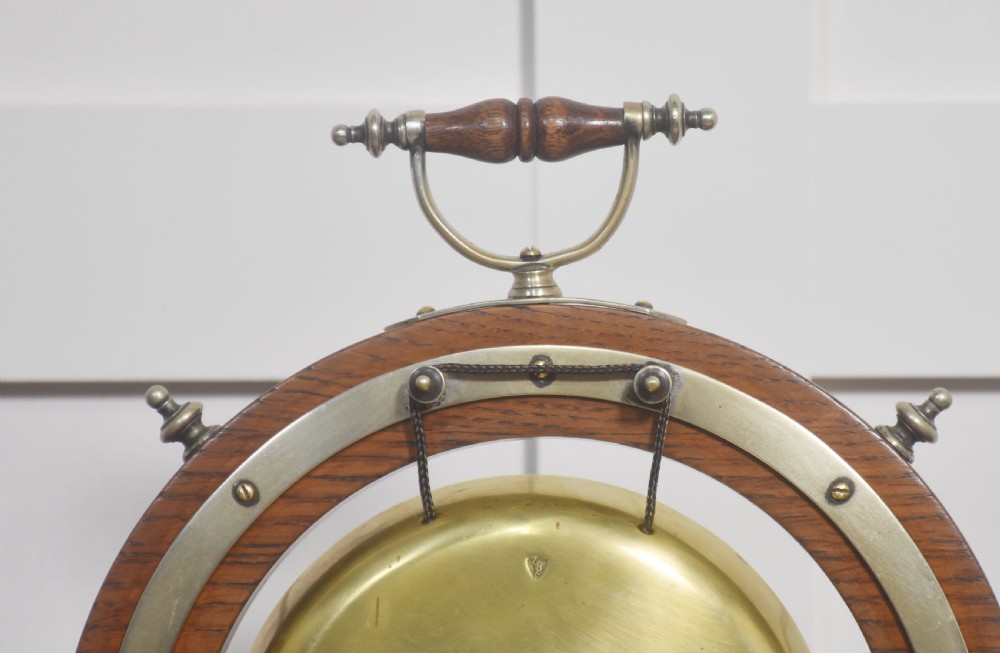 An antique oak ship wheel frame holding a central suspended brass dinner gong with an accompanying wooden striker resting on brass brackets on a rectangular base. View IV