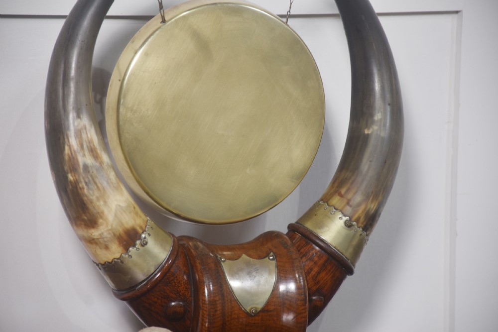 An antique tabletop dinner gong featuring a circular brass disc suspended by chains between two large curved cattle horns on a carved oak base. View IV