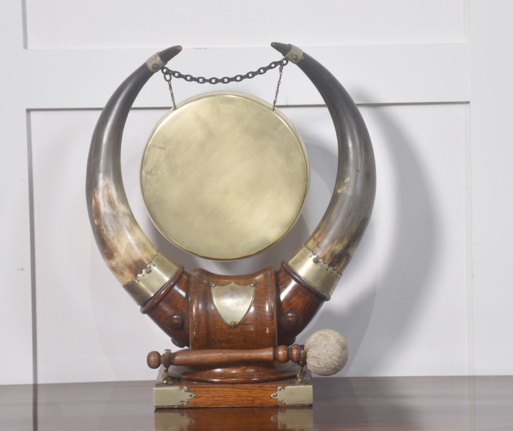 An antique tabletop dinner gong featuring a circular brass disc suspended by chains between two large curved cattle horns on a carved oak base. View V