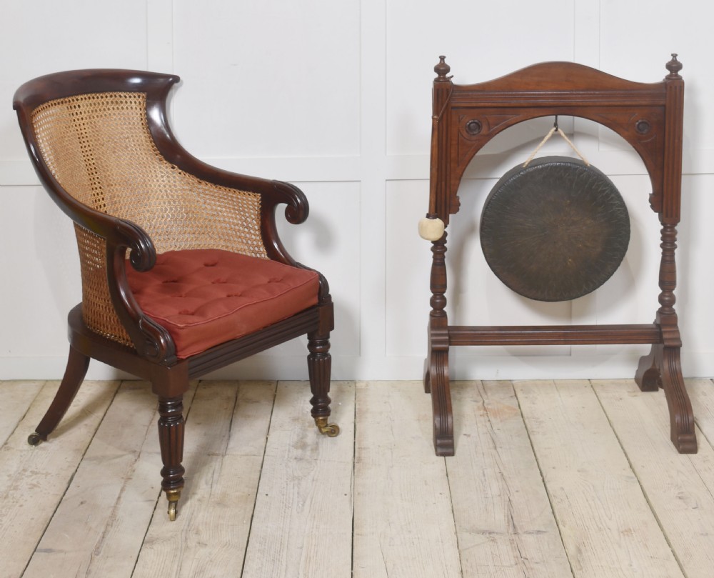 A Regency mahogany bergere armchair featuring a curved woven cane back, scrolled armrests, a buttoned terracotta squab cushion, and turned front legs on brass castors. View II