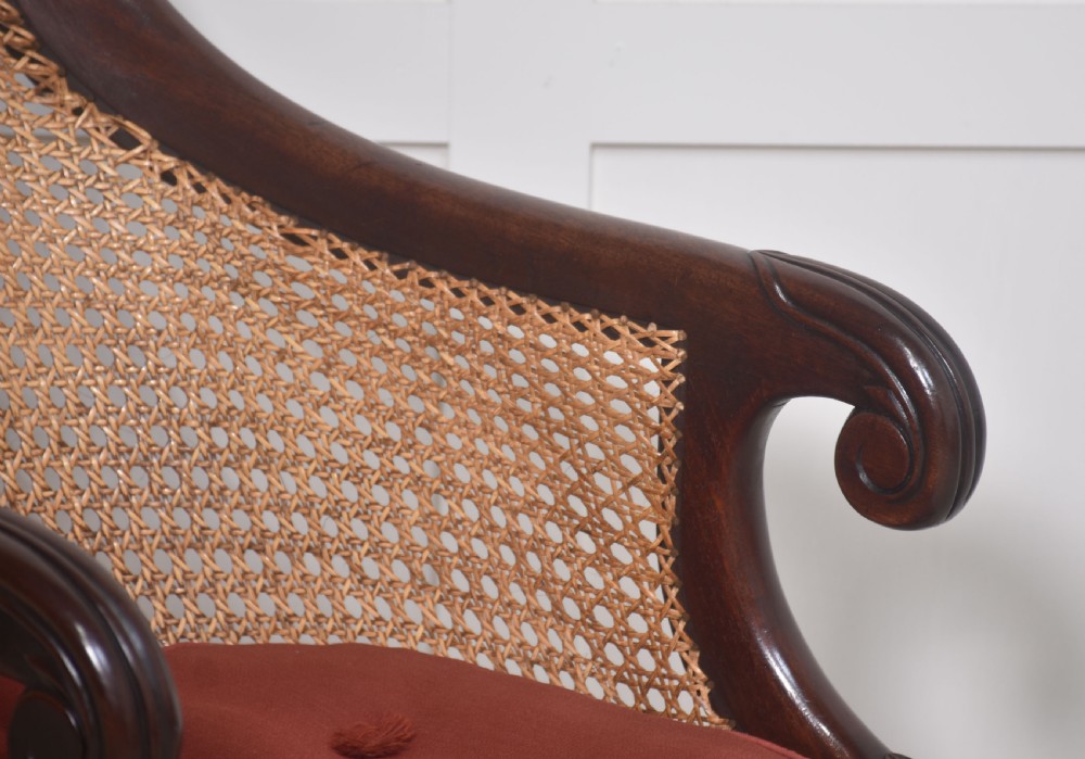A Regency mahogany bergere armchair featuring a curved woven cane back, scrolled armrests, a buttoned terracotta squab cushion, and turned front legs on brass castors. View III