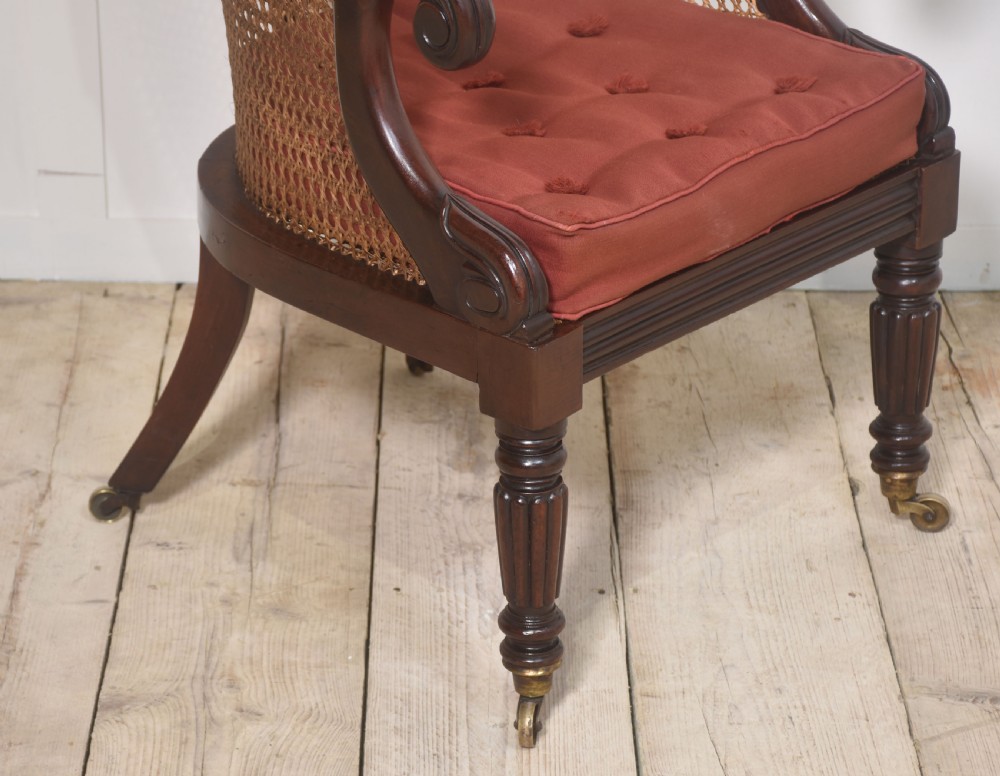 A Regency mahogany bergere armchair featuring a curved woven cane back, scrolled armrests, a buttoned terracotta squab cushion, and turned front legs on brass castors. View VII