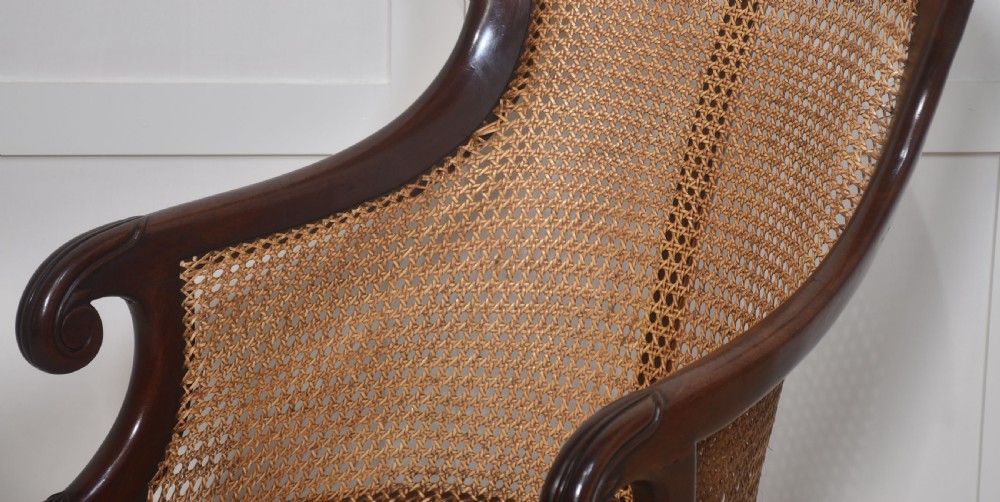 A Regency mahogany bergere armchair featuring a curved woven cane back, scrolled armrests, a buttoned terracotta squab cushion, and turned front legs on brass castors. View X