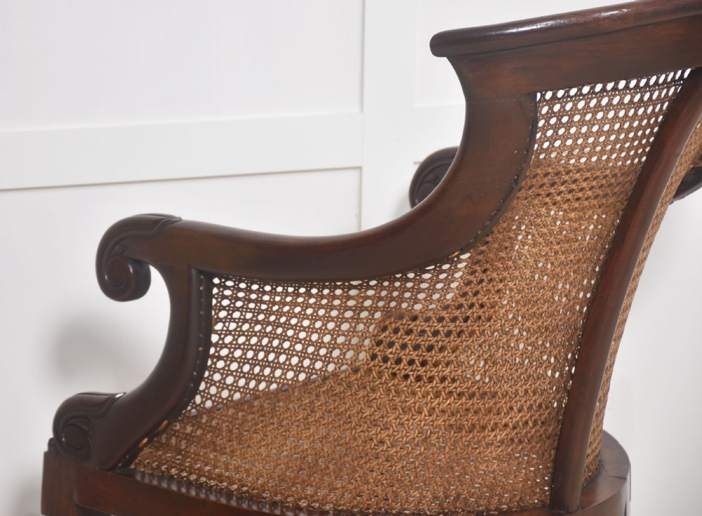 A Regency mahogany bergere armchair featuring a curved woven cane back, scrolled armrests, a buttoned terracotta squab cushion, and turned front legs on brass castors. View XII