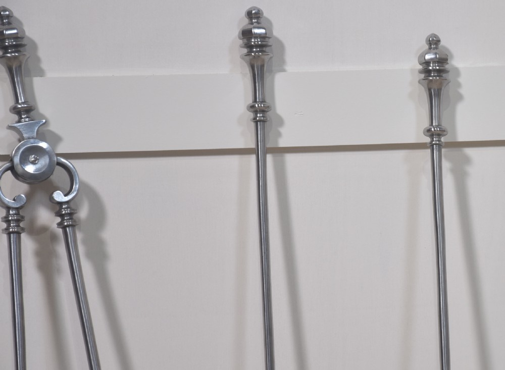 A set of three polished steel fire tools including tongs, a pierced shovel, and a poker with turned finial handles standing upright. View IV