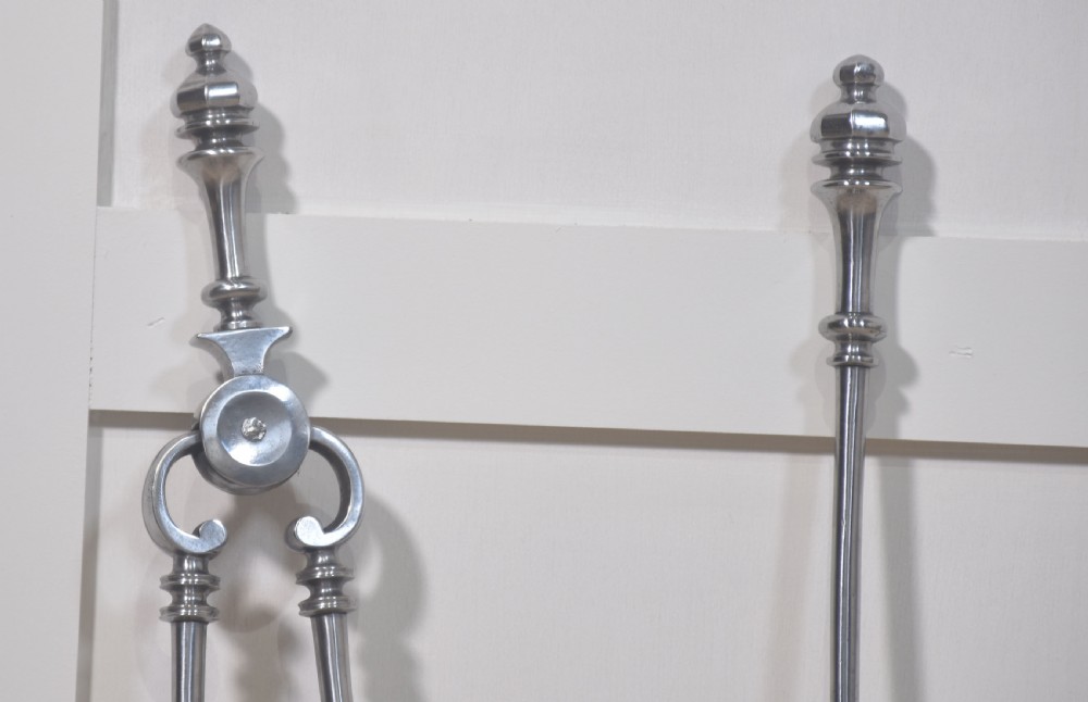 A set of three polished steel fire tools including tongs, a pierced shovel, and a poker with turned finial handles standing upright. View V