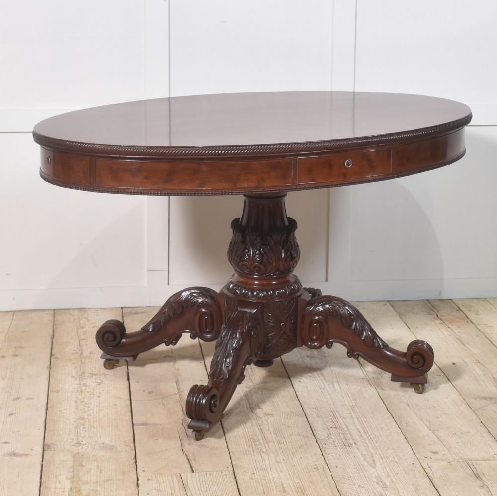 A 19th-century oval mahogany drum table featuring a reeded top edge, frieze drawers, and an elaborately carved acanthus pedestal on scroll legs with brass castors.