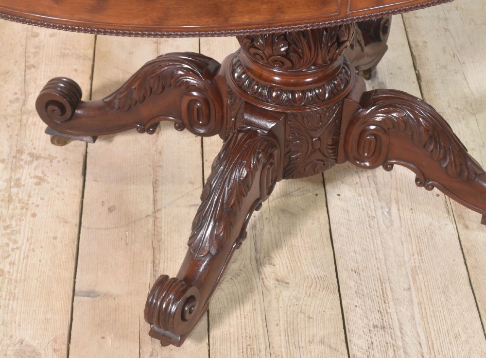 A 19th-century oval mahogany drum table featuring a reeded top edge, frieze drawers, and an elaborately carved acanthus pedestal on scroll legs with brass castors. View VI