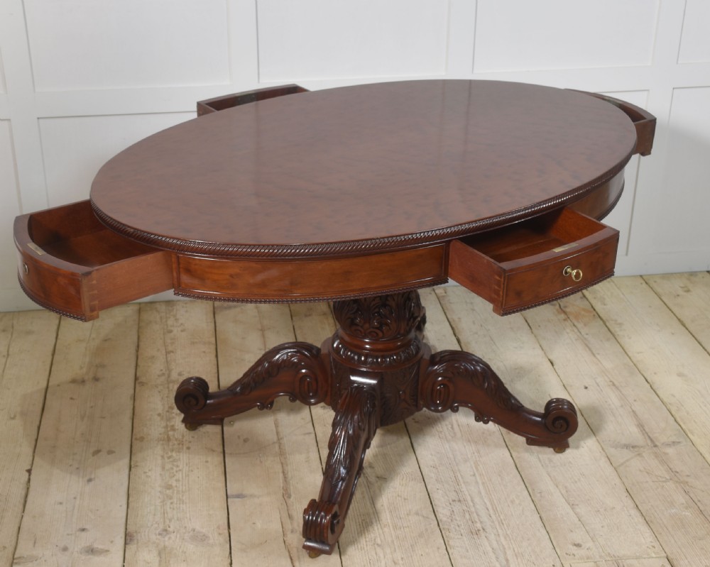 A 19th-century oval mahogany drum table featuring a reeded top edge, frieze drawers, and an elaborately carved acanthus pedestal on scroll legs with brass castors. View X
