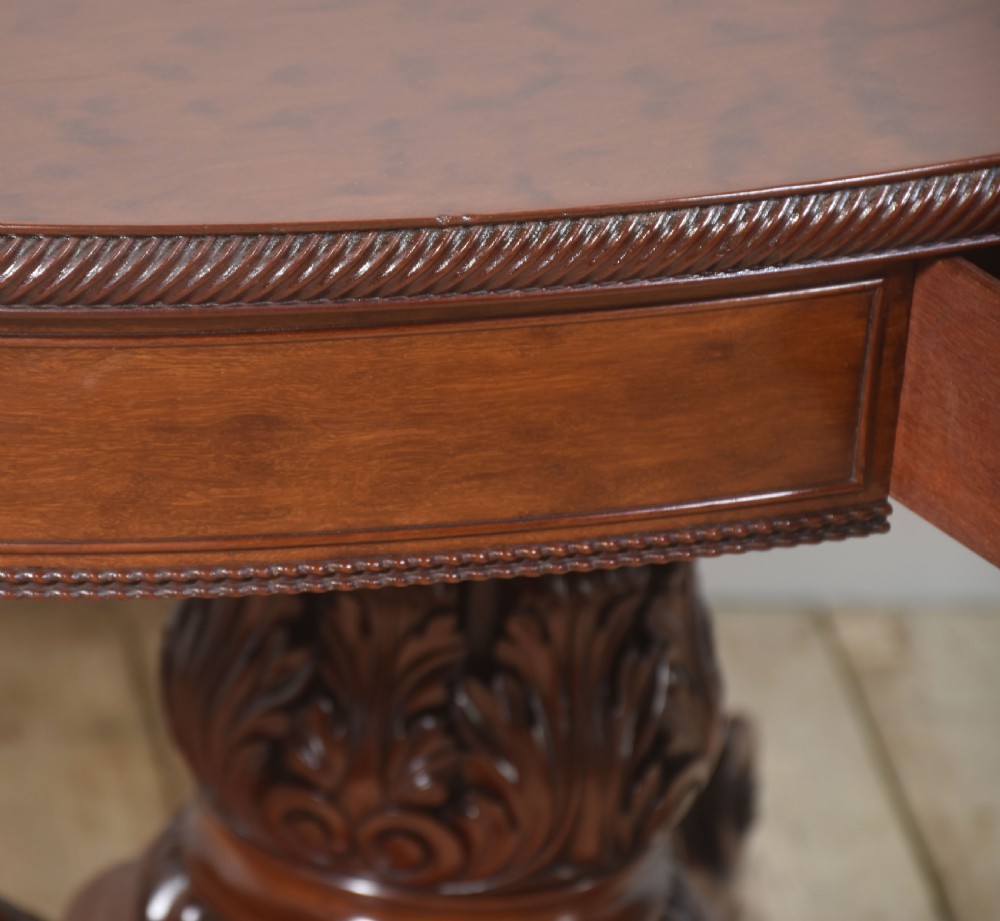 A 19th-century oval mahogany drum table featuring a reeded top edge, frieze drawers, and an elaborately carved acanthus pedestal on scroll legs with brass castors. View XII