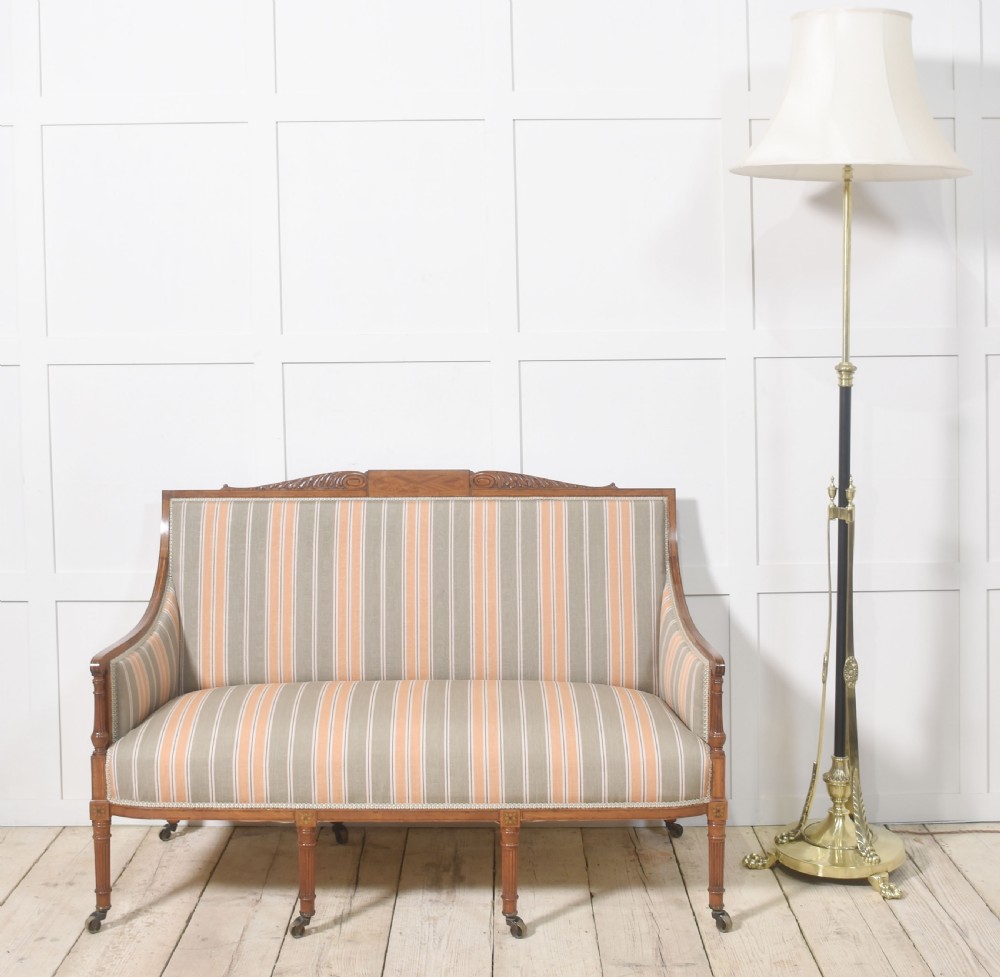 An Edwardian satinwood inlaid two-seater settee with striped orange and grey fabric upholstery, resting on turned front legs with brass castors. View II