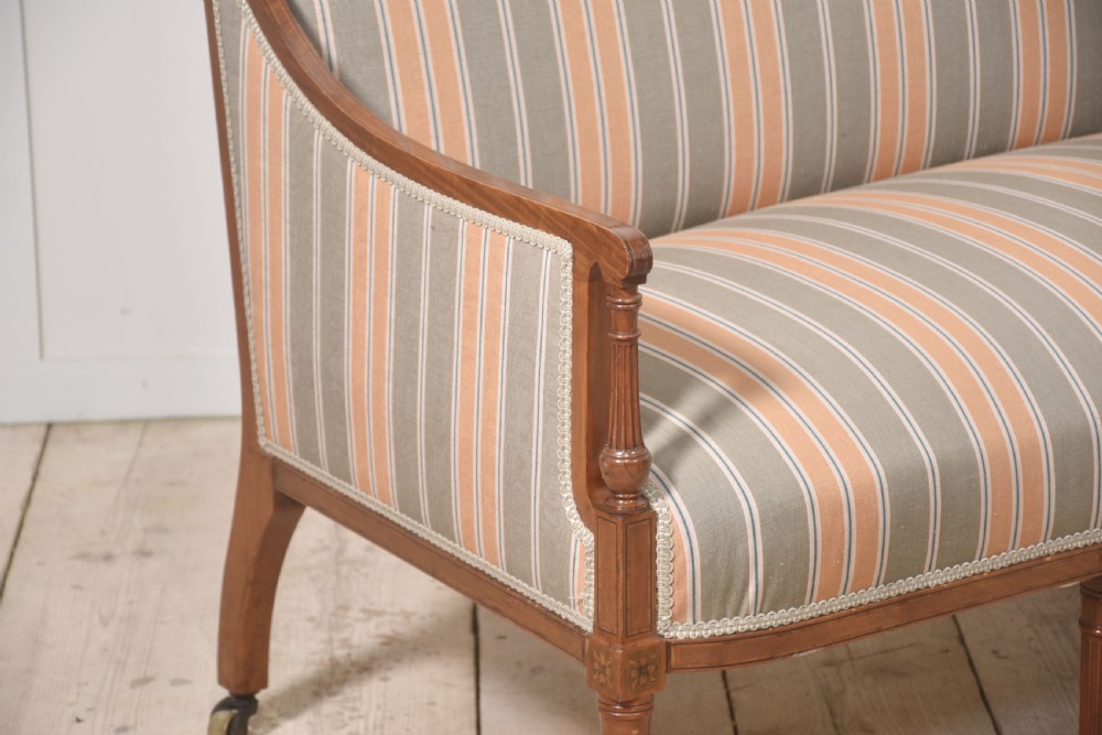 An Edwardian satinwood inlaid two-seater settee with striped orange and grey fabric upholstery, resting on turned front legs with brass castors. View VII