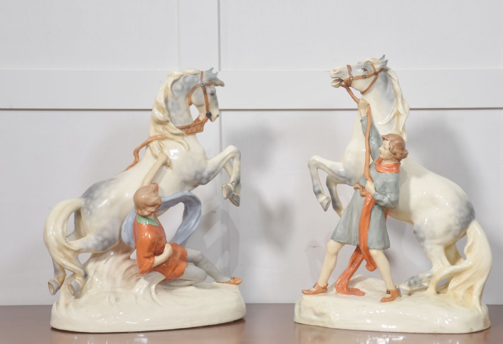 A pair of glazed cream and grey porcelain figures depicting rearing horses held by handlers on naturalistic bases.