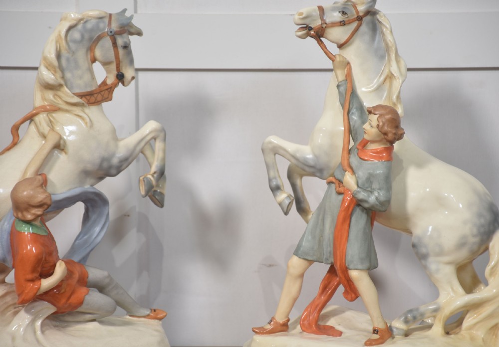 A pair of glazed cream and grey porcelain figures depicting rearing horses held by handlers on naturalistic bases. View V