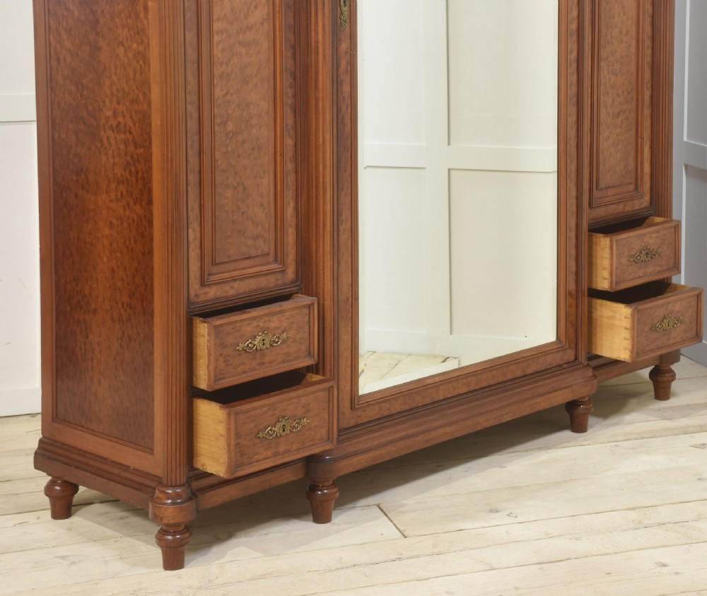 A French Louis XVI style mahogany triple armoire featuring a central mirrored door flanked by carved timber panels and lower drawers. View XVII