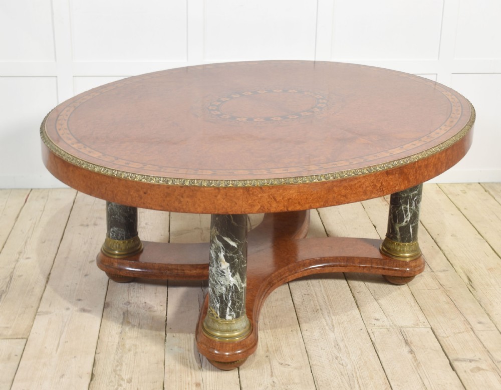 An oval centre table featuring a burr walnut marquetry top, brass rim moulding, four green marble column supports, and a curved stretcher base.